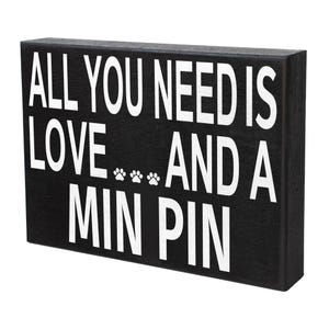 May include: Black wooden sign with white text that reads "All you need is love... and a Min Pin". There are three paw prints between the words "love" and "and".