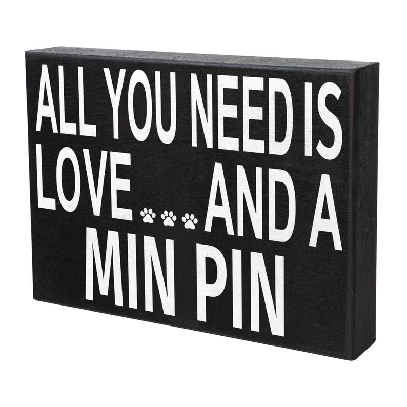 May include: Black wooden sign with white text that reads "All you need is love... and a Min Pin". There are three paw prints between the words "love" and "and".