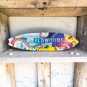 May include: A surfboard-shaped wooden sign with the word "Wildwoods" in blue and white lettering. The sign features a colorful illustration of an amusement park with a Ferris wheel, roller coaster, and other attractions. The sign is displayed on a wooden shelf.