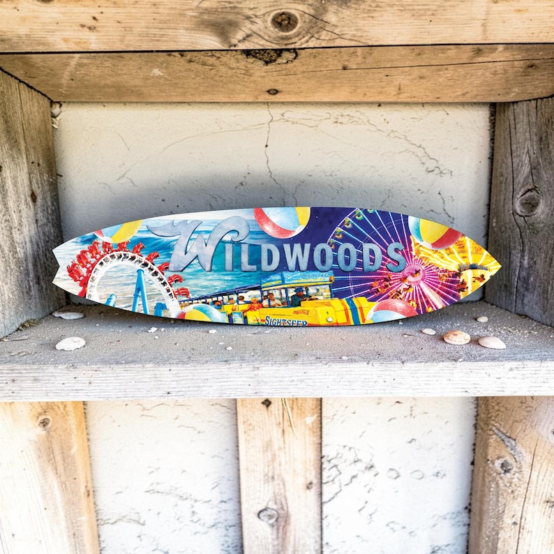 May include: A surfboard-shaped wooden sign with the word "Wildwoods" in blue and white lettering. The sign features a colorful illustration of an amusement park with a Ferris wheel, roller coaster, and other attractions. The sign is displayed on a wooden shelf.