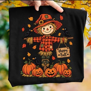Könnte beinhalten: Ein schwarzes T-Shirt mit einer Grafik einer Vogelscheuche, die ein rot-schwarz kariertes Hemd und einen Hut mit Sonnenblume trägt. Die Vogelscheuche hält ein Holzschild mit der Aufschrift Happy Fall. Schwarze Krähen und Kürbisse umgeben sie.