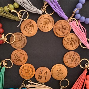 May include: A collection of wooden keychains with various phrases and designs, including "Wake Pray Slay", "Blessed Mama", "Kinda Classy Kinda Savage", "Not Today Satan", "You Tough But So Are Babe", "I Love", and "It Was Like Ten Minutes Ago Friday". Each keychain is attached to a beaded chain with a tassel.