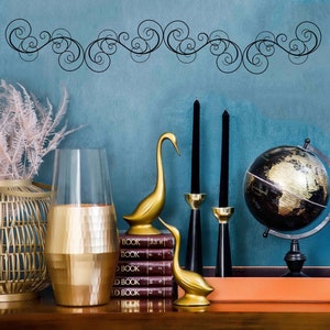 Puede incluir: Un jarrón decorativo dorado con acabado de metal martillado, una cesta de mimbre con flores rosas secas, dos figuras de cisnes dorados, un globo terráqueo negro y dos candelabros negros con velas negras sobre una mesa de madera.