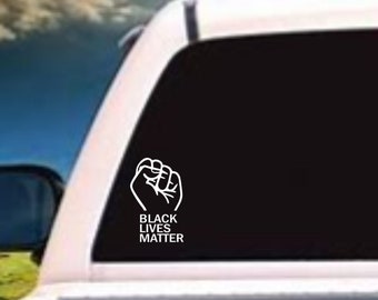 May include: Black Lives Matter decal with a raised fist graphic on a black background. The decal is on the back window of a white pickup truck.
