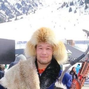 Può includere: Un uomo con un cappello di pelliccia tradizionale e una tunica blu tiene un'aquila reale, una pelle di lupo e un faretra di frecce. È in piedi su una piattaforma di legno con montagne innevate sullo sfondo.