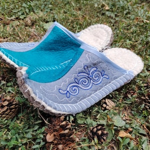 May include: A pair of felt slippers with a teal and gray design, accented with white stitching and blue embroidered swirls. The slippers have a light-colored, fluffy interior and are set on a bed of green grass and pine cones.