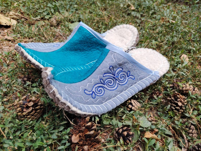 May include: A pair of felt slippers with a teal and gray design, accented with white stitching and blue embroidered swirls. The slippers have a light-colored, fluffy interior and are set on a bed of green grass and pine cones.
