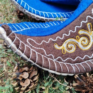 May include: Close-up of a pair of felt boots. The boots are blue and brown with white and yellow embroidered details. The boots are decorated with intricate patterns and stitching. The boots are on a bed of grass and pine cones.