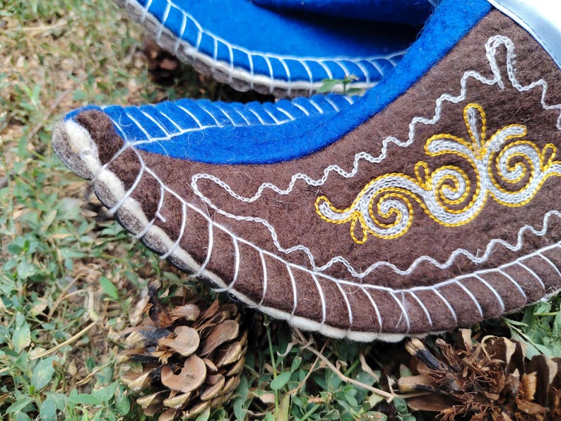 May include: Close-up of a pair of felt boots. The boots are blue and brown with white and yellow embroidered details. The boots are decorated with intricate patterns and stitching. The boots are on a bed of grass and pine cones.
