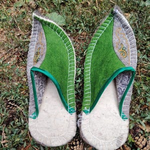 May include: A pair of handmade felt slippers. The slippers have a light gray base with a green top and a decorative embroidered design. The edges are trimmed with white stitching. The slippers are designed to be worn indoors.