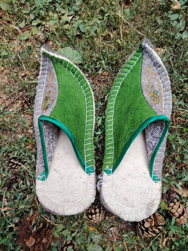 May include: A pair of handmade felt slippers. The slippers have a light gray base with a green top and a decorative embroidered design. The edges are trimmed with white stitching. The slippers are designed to be worn indoors.