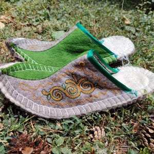 May include: A pair of handmade felt slippers with a gray base, green accents, and intricate gold embroidery. The slippers have a white stitched border and a green trim. The design includes a leaf-like shape and a decorative swirl pattern.