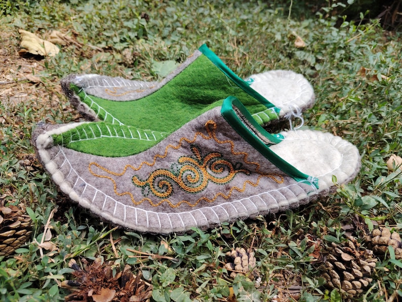 May include: A pair of handmade felt slippers with a gray base, green accents, and intricate gold embroidery. The slippers have a white stitched border and a green trim. The design includes a leaf-like shape and a decorative swirl pattern.