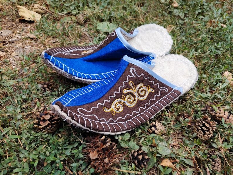 May include: A pair of handmade felt slippers with a brown and blue design. The slippers feature white stitching and embroidered gold and yellow accents. The interior is lined with white, fluffy material. The slippers are displayed on a bed of green grass and pine cones.