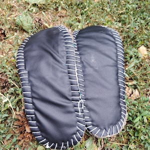 May include: A pair of black leather slippers with white stitching around the edges. The slippers are resting on a bed of green grass and fallen leaves, showcasing a handcrafted design.