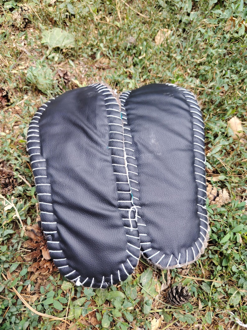 May include: A pair of black leather slippers with white stitching around the edges. The slippers are resting on a bed of green grass and fallen leaves, showcasing a handcrafted design.