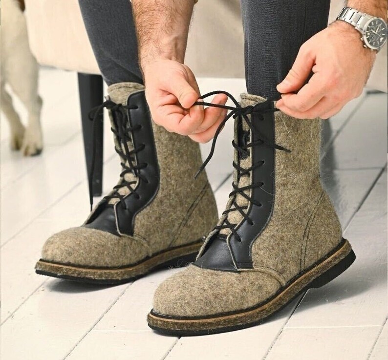 May include: A person tying the laces of a pair of tall, gray felt boots with black leather accents. The boots have a lace-up design and a sturdy sole. The boots are a neutral color, suitable for various outfits.