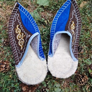 May include: A pair of handmade felt slippers. The slippers feature a white sole, a brown and blue upper with decorative stitching, and a paisley patterned interior. The slippers are displayed on a bed of green grass and pine needles.