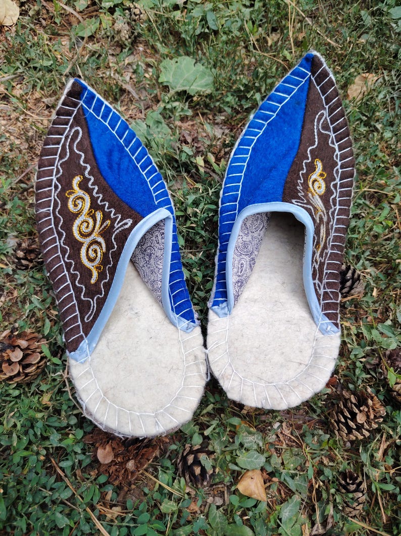 May include: A pair of handmade felt slippers. The slippers feature a white sole, a brown and blue upper with decorative stitching, and a paisley patterned interior. The slippers are displayed on a bed of green grass and pine needles.