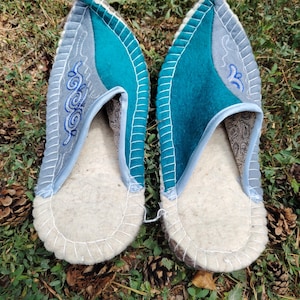 May include: A pair of felt slippers with a light gray and teal color scheme. The slippers have a white sole and are decorated with embroidered swirls. The slippers are on a bed of green grass and pine cones.