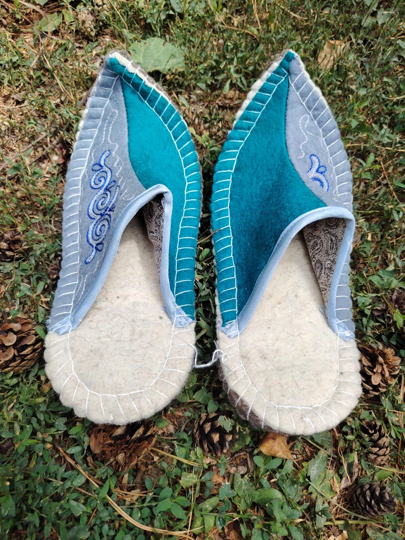 May include: A pair of felt slippers with a light gray and teal color scheme. The slippers have a white sole and are decorated with embroidered swirls. The slippers are on a bed of green grass and pine cones.