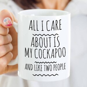 May include: White ceramic mug with the text "ALL I CARE ABOUT IS MY COCKAPOO AND LIKE TWO PEOPLE" in black, wavy lines above and below the text. The mug is held by a person.