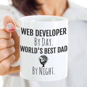 May include: White ceramic mug with black text that reads "WEB DEVELOPER BY DAY. WORLD'S BEST DAD BY NIGHT." A small globe graphic is below "WORLD'S BEST DAD." The mug is held by a person.