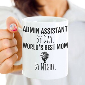 May include: White ceramic coffee mug with black text that reads "Admin Assistant by Day. World's Best Mom by Night."  A small globe graphic is between the two lines of text.