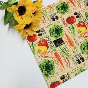 May include: A beige kitchen towel with a colorful vegetable print, including tomatoes, carrots, lettuce, and olives. The towel also features the text "Market Fresh". A bouquet of yellow sunflowers rests on the corner of the towel.