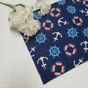 May include: A navy blue nautical-themed placemat featuring a pattern of white anchors, light blue ship wheels, and red and white life preservers. White flowers are placed on the corner of the placemat. The placemat is made of fabric.