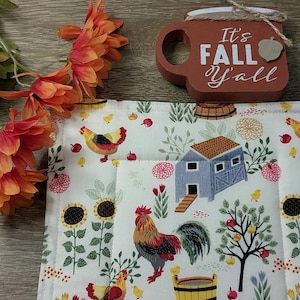 May include: A white quilted placemat with a farm scene featuring chickens, sunflowers, and a blue barn. A wooden sign that says "It's FALL Y'all" sits above the placemat. Orange flowers are in the upper left corner.