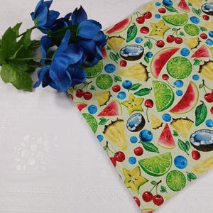 May include: A light green table runner with a colorful fruit pattern featuring watermelon, pineapple, cherries, blueberries, star fruit, coconut, and lime slices. Blue artificial flowers are on the left side of the image.