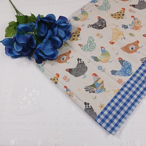 May include: A beige quilted placemat featuring a repeating pattern of colorful chickens. The placemat has a blue and white gingham border. A bouquet of blue flowers rests on the white tablecloth.