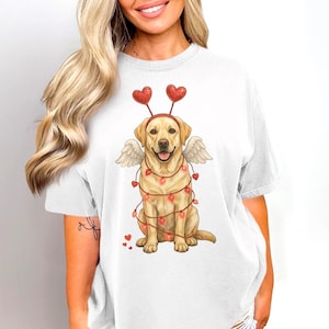 May include: White t-shirt featuring a cartoon Labrador Retriever dressed as Cupid for Valentine's Day. The dog has heart-shaped antennae, angel wings, and is wrapped in a string of heart-shaped lights. The design includes small red hearts.