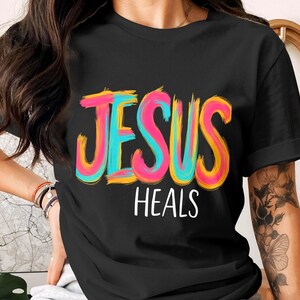 Può includere: T-shirt nera con la scritta "GESÙ GUARISCE" in lettere colorate. La parola "GESÙ" è in blu, rosa e giallo. "GUARISCE" è bianca. La maglietta ha maniche corte e scollo tondo.