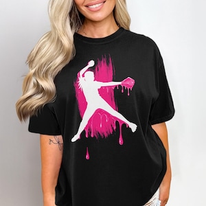 May include: Black t-shirt featuring a white silhouette of a softball player in action, set against a vibrant pink paint stroke design. The design includes dripping paint effects, creating a dynamic and eye-catching graphic.
