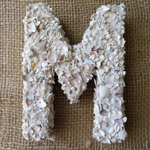May include: A white letter M made of seashells. The letter is on a burlap background.