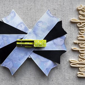 May include: A handmade hair bow with a layered design. The bow features blue and white striped fabric, black fabric, and a yellow and green accent. The word "bekadetpauses" is written in a cursive font.