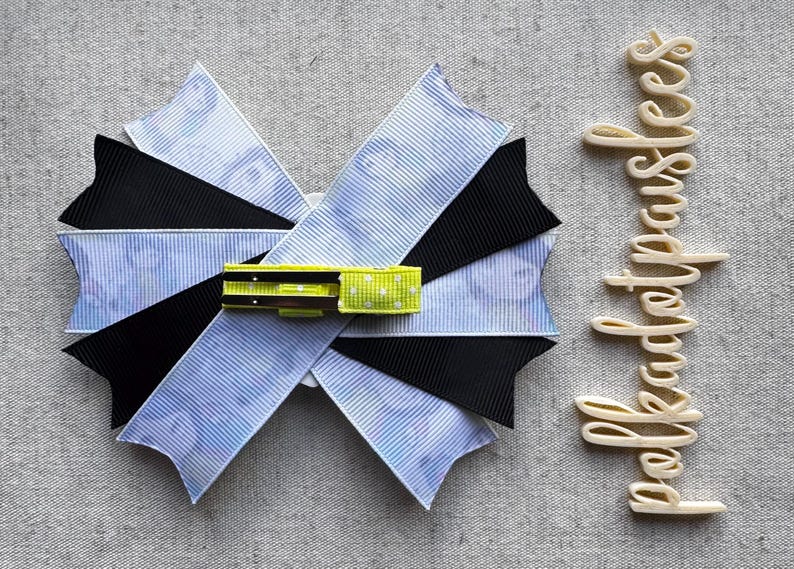 May include: A handmade hair bow with a layered design. The bow features blue and white striped fabric, black fabric, and a yellow and green accent. The word "bekadetpauses" is written in a cursive font.