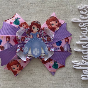 May include: A large, layered hair bow featuring a cartoon princess. The bow is made of purple and pink ribbons with princess graphics. A 3D princess figure is centered on the bow. The bow is a decorative hair accessory.