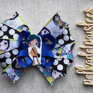 May include: A handmade hair bow featuring a cartoon character design. The bow is made of layered ribbons in black with white polka dots, and a patterned ribbon with a cartoon character and spiderwebs. A blue ribbon is in the center.