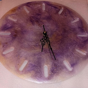 May include: Round wall clock with a marbled design in shades of purple, gold, and white. The clock face has white rectangular hour markers and black clock hands. A decorative piece for home decor.