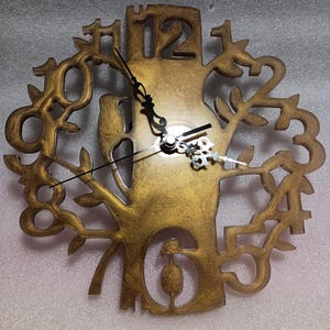 May include: A gold-colored, tree-themed wall clock with cut-out numbers and a bird silhouette. The clock hands are black, and the clock face is a warm, metallic gold. The design incorporates a tree trunk and branches with the numbers integrated into the design.