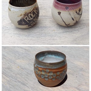 Puede incluir: Tres tazas de cerámica hechas a mano. Una taza es beige con la palabra "ROCK" en marrón oscuro. Otra es beige con un borde granate y diseños florales. La tercera taza es marrón con patrones de puntos azules y verdes.