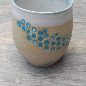 Puede incluir: Una taza de cerámica blanca con un glaseado azul claro en el borde y un patrón de puntos azules en el cuerpo beige.