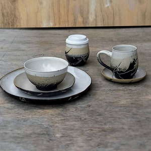 Puede incluir: Un juego de vajilla de cerámica con un diseño en blanco y negro. El juego incluye un tazón, un plato, una taza y una taza con tapa. Todos los platos están decorados con una imagen estilizada de un pez.