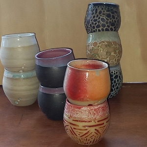 May include: A set of six ceramic cups, stacked in two groups of three. The cups are all different colors and patterns, including black, white, brown, red, and orange. The cups are all hand-crafted and have a unique, rustic look.