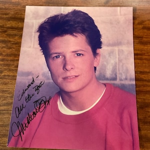 Puede incluir: Una fotografía en blanco y negro de un joven que lleva una sudadera rosa. El hombre está mirando a la cámara y sonriendo ligeramente. La foto está firmada "Edward - All the best - Michael J. Fox".