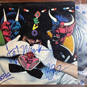 以下が含まれることがあります： 2匹の角のある悪魔のような人物と白い手袋をした人物のカラフルなイラストが描かれた、ヴィンテージのビニールレコードアルバムのカバー。アルバムカバーには、バンド名「DEVO」が緑と紫で表示されています。カバーにはバンドメンバーのサインが入っています。