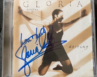 May include: A signed CD by Gloria Estefan, titled "Destiny". The cover features a black and white photo of Gloria Estefan in a black dress, with the title "Gloria Estefan" and "Destiny" printed in white.